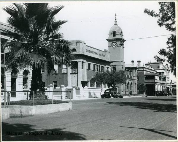 The post office in 1930. Palm trees in raised beds were a feature of the main street. The palms were removed in 1945. A car and motorcycle with side car are parked outside the Government Savings Bank. Image Macquarie Regional Library.